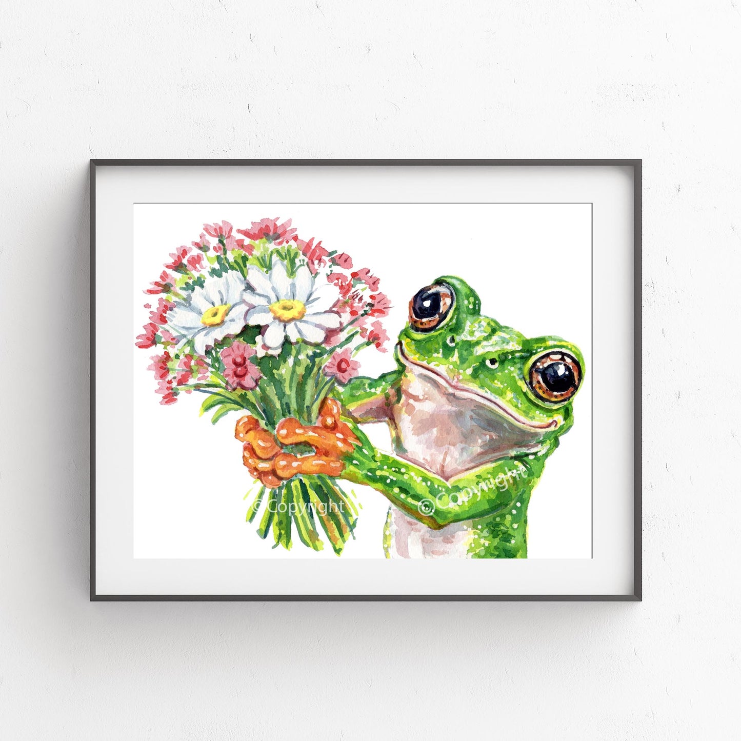 A framed watercolor art print of a green tree frog holding a bouquet of white and pink flowers, mounted on a white textured wall.