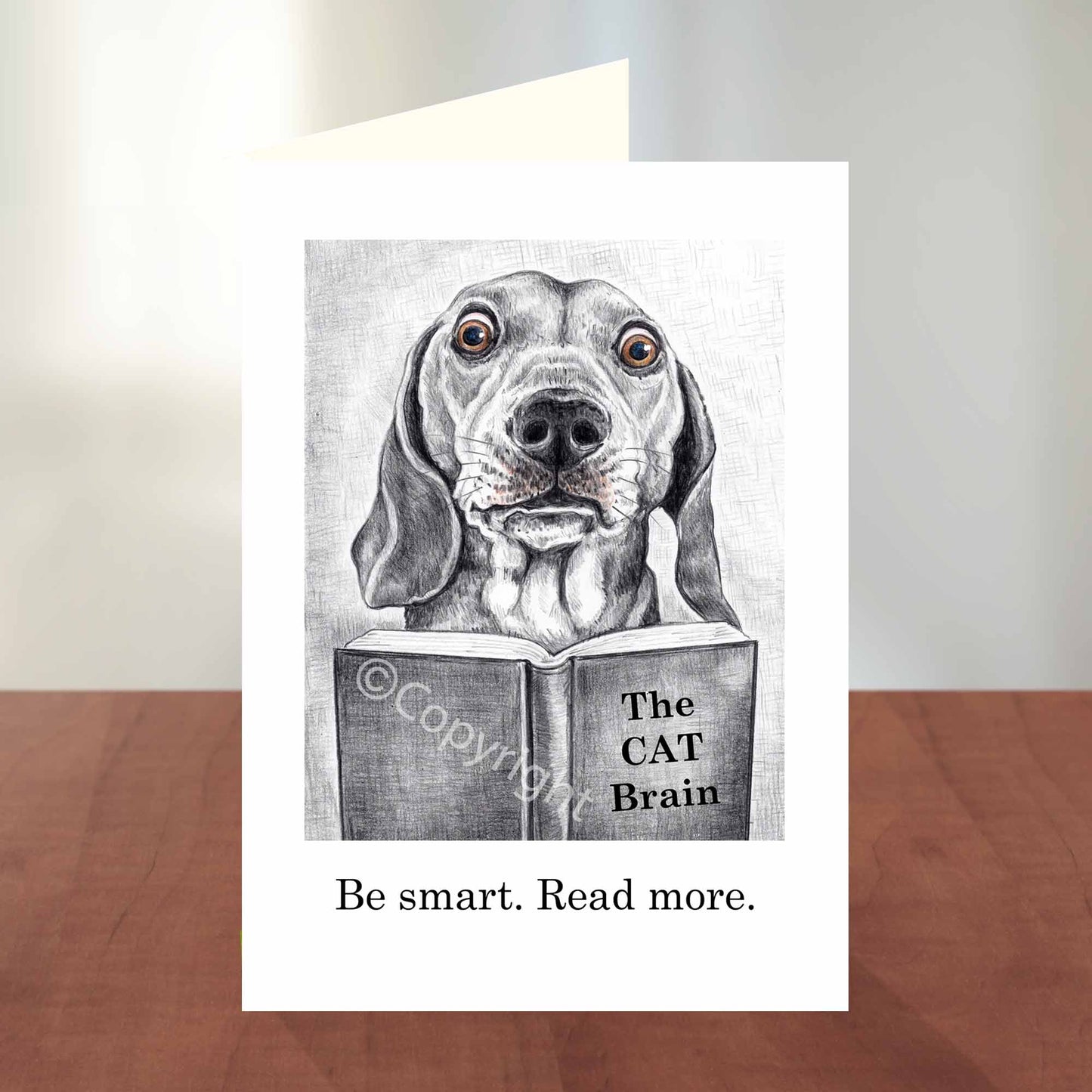 Greeting card featuring a wax pastel drawing of a dachshund dog reading a book about cats. Black and white with a hint of colour. By Deidre Wicks