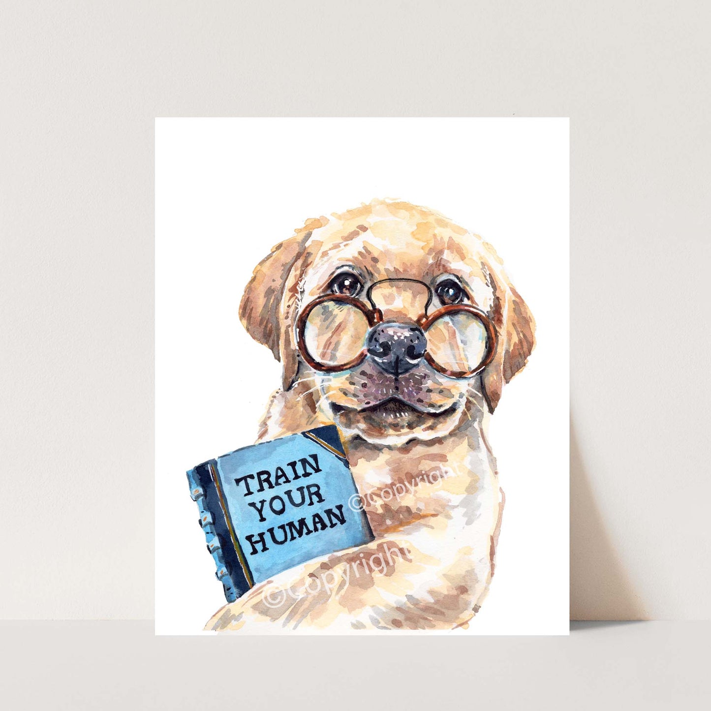 Yellow lab puppy dog wearing glasses holding a book titled 'Train Your Human' on a white background