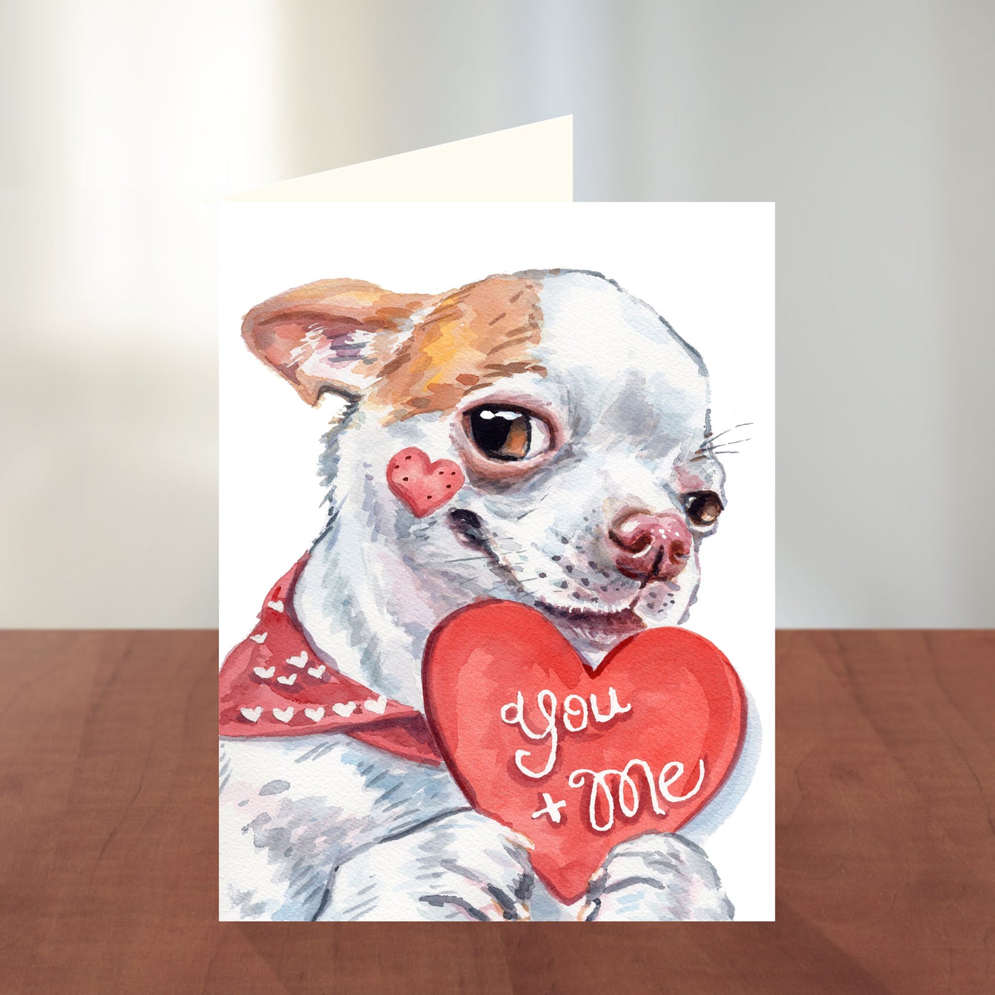 Greeting card with a dog holding a heart that says 'you + me' on a wooden surface.