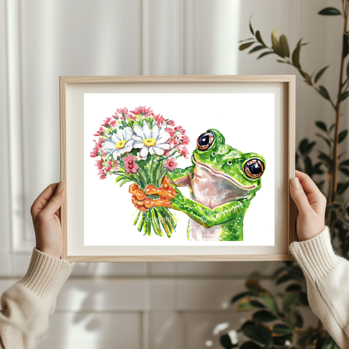 A person holds a framed watercolor painting of a green tree frog holding a bouquet of white, pink, and yellow flowers. The scene is set in a bright, modern room with a white paneled wall and a green plant in the background.