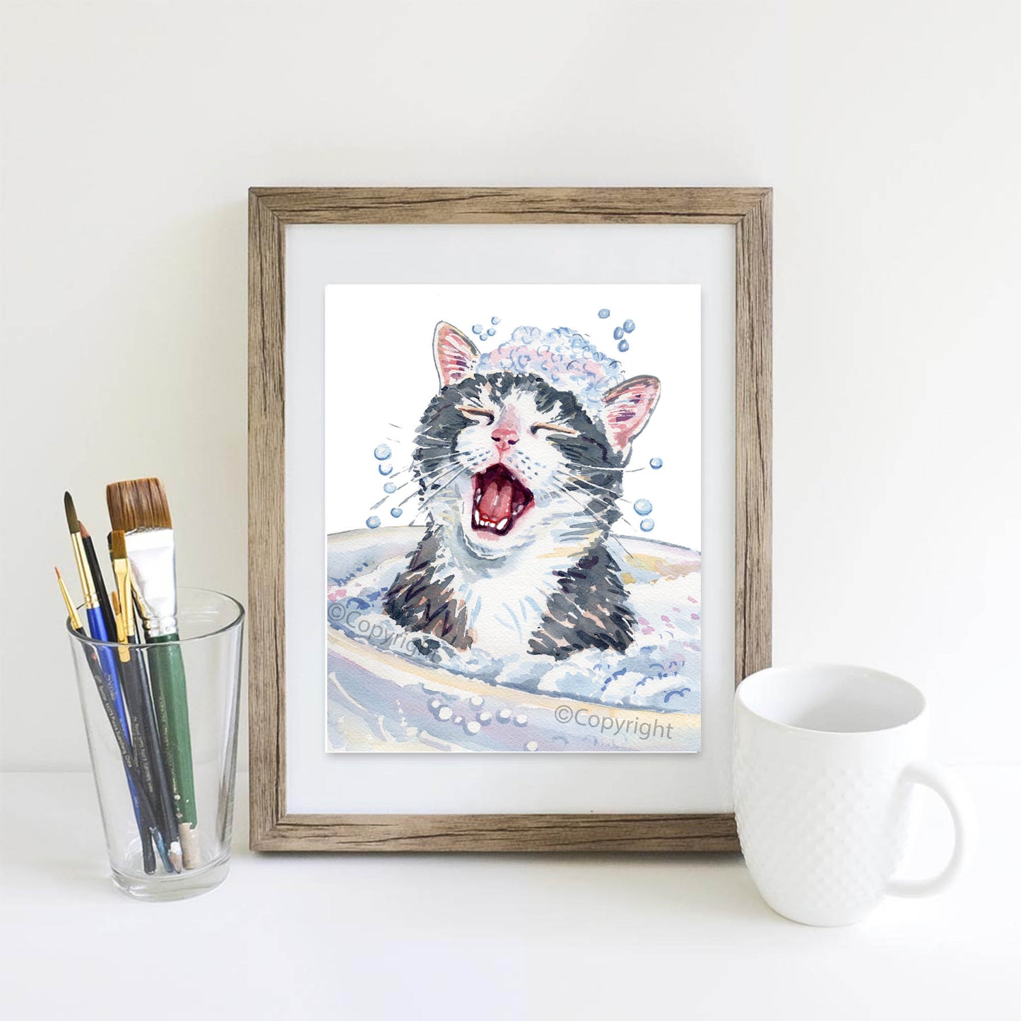 Framed illustration of a cat in a bath with art supplies and a mug on a white surface