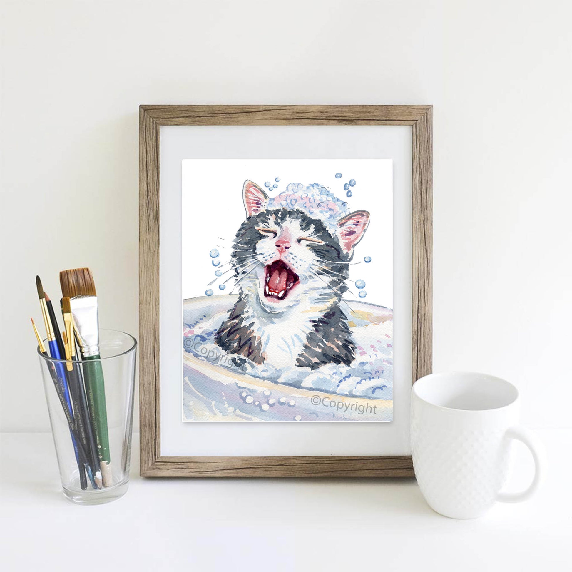 Framed illustration of a cat in a bath with art supplies and a mug on a white surface