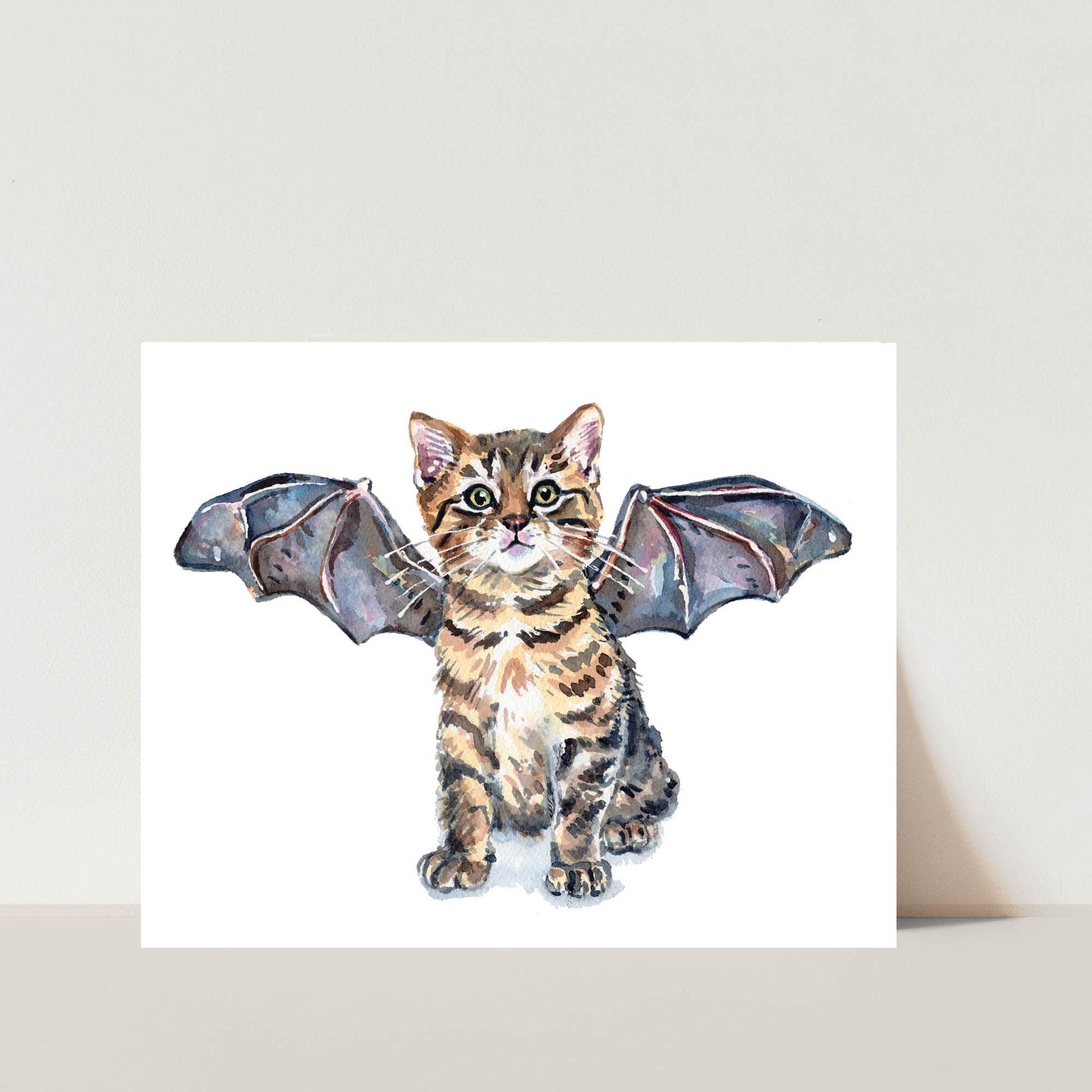 A watercolor art print featuring a small tabby kitten with wings spread, shown against a white square print mounted on a light beige wall.
