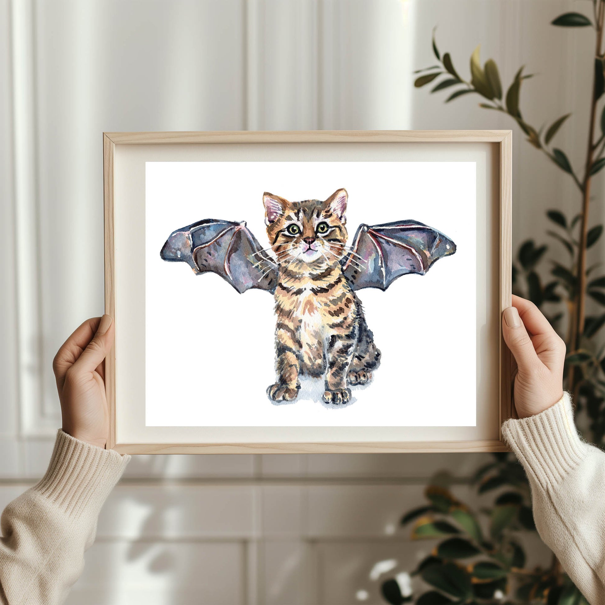 A person holds a light wood framed art print featuring a watercolor illustration of a tabby kitten with bat wings. The person is wearing cream-colored knit sleeves, and a blurred indoor background with a plant is visible.