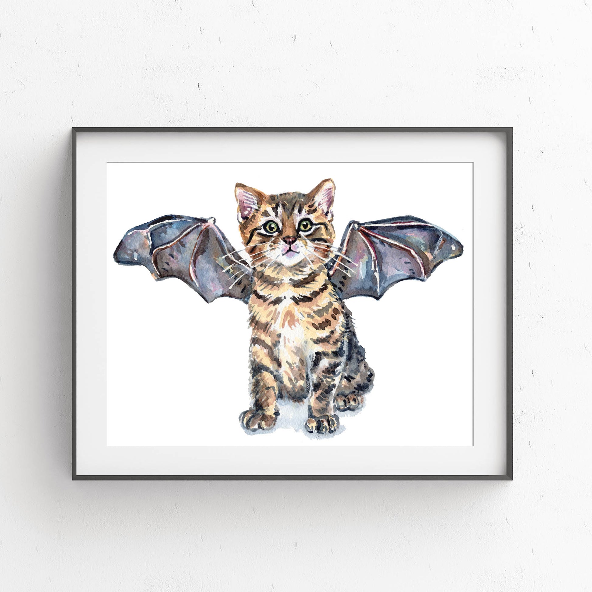 A framed watercolor art print of a tabby kitten with wings spread, shown against a white wall. The print features a white background with a detailed illustration of the kitten in brown and black fur, wearing a bat costume.