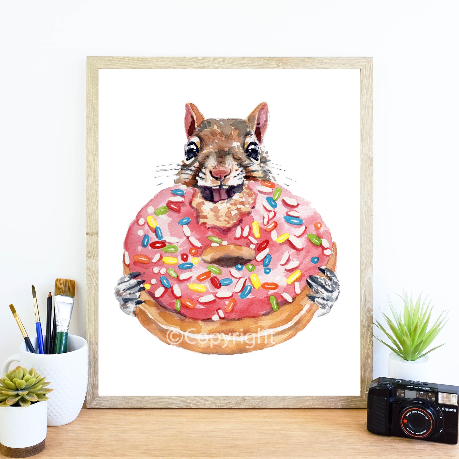 Framed artwork of a squirrel holding a pink donut with sprinkles on a desk.