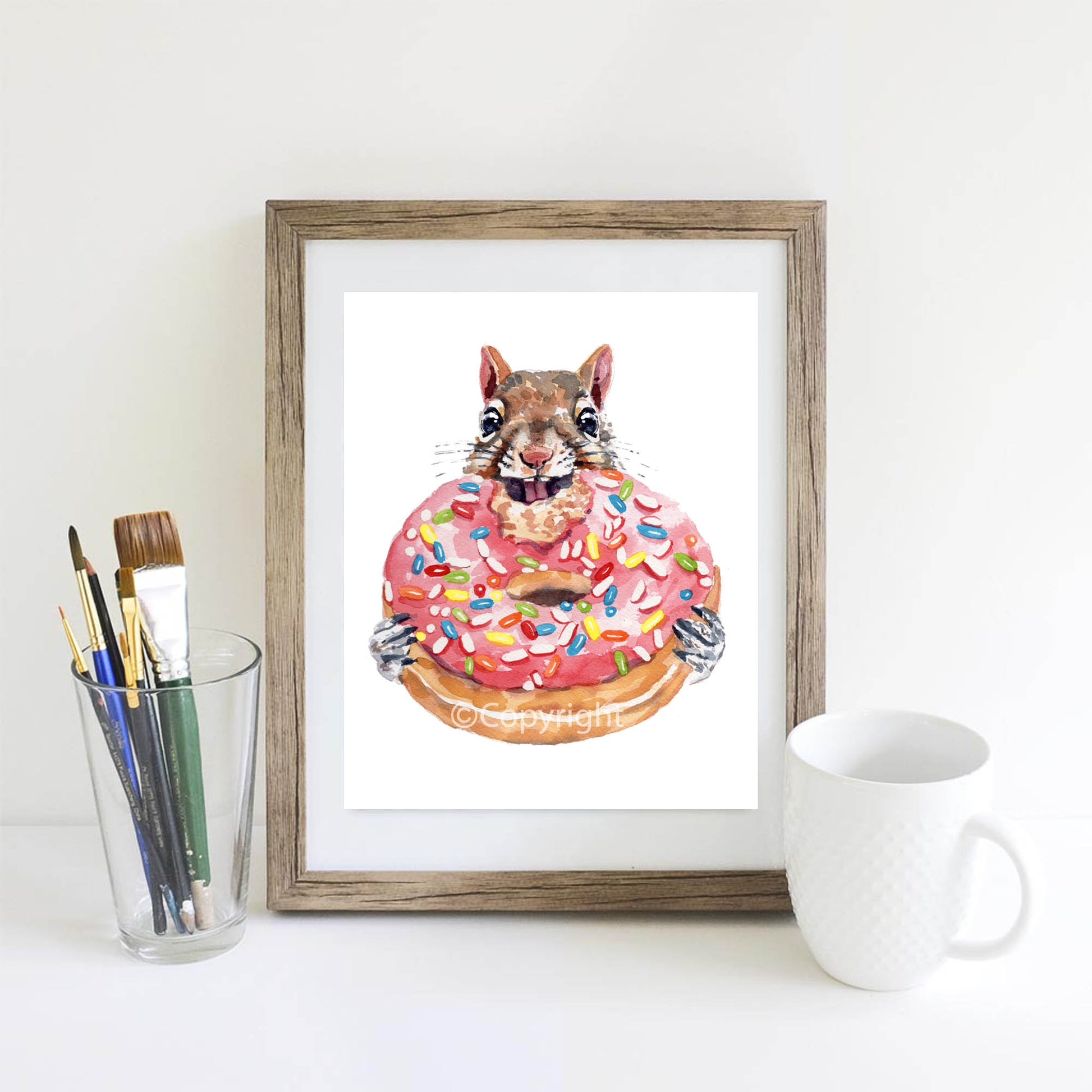 Framed artwork of a squirrel on a donut with colorful sprinkles, placed on a white surface with art supplies and a mug.