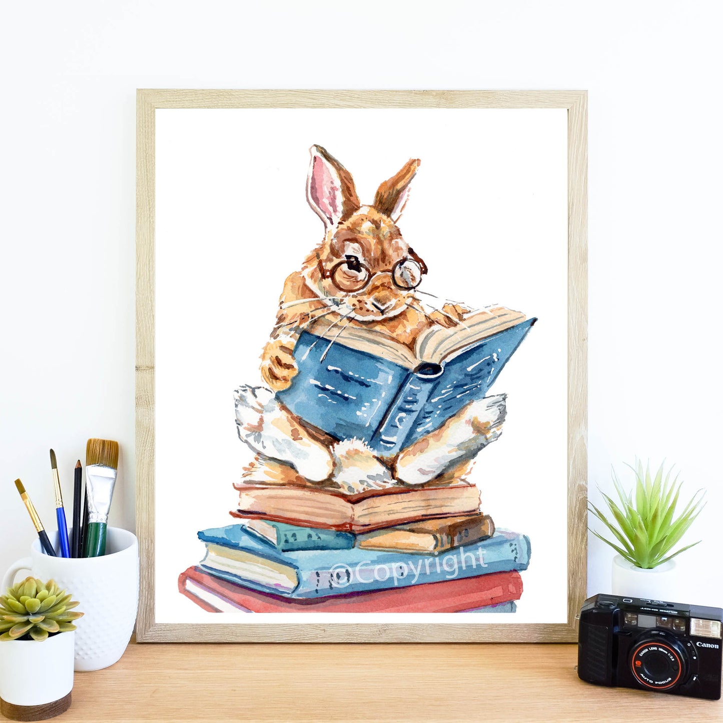 Framed artwork of a rabbit reading a book on a stack of books with stationery items on a desk.
