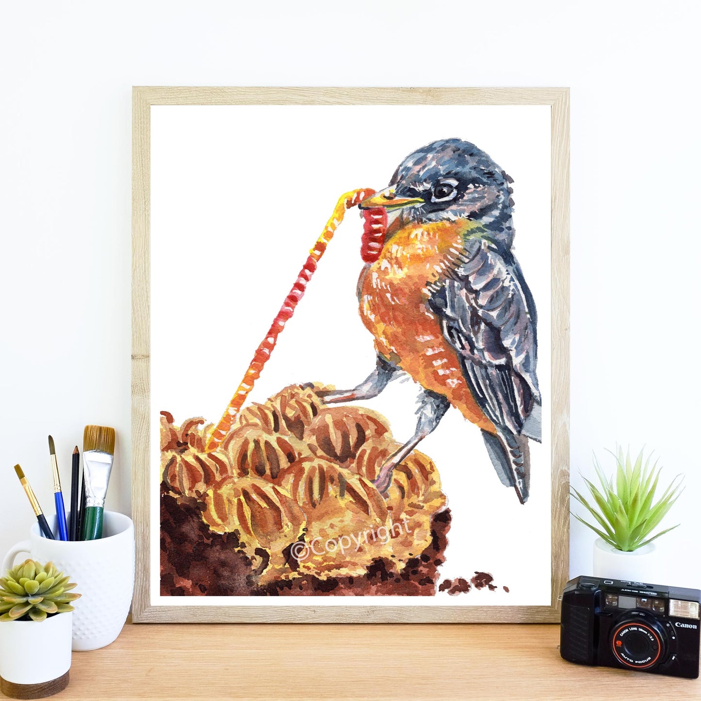 Framed artwork of a bird on a cake with stationery items and a camera on a desk.