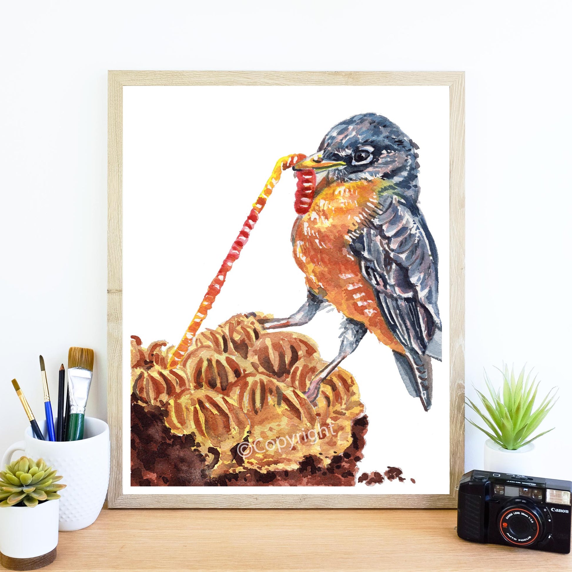Framed artwork of a bird on a cake with stationery items and a camera on a desk.