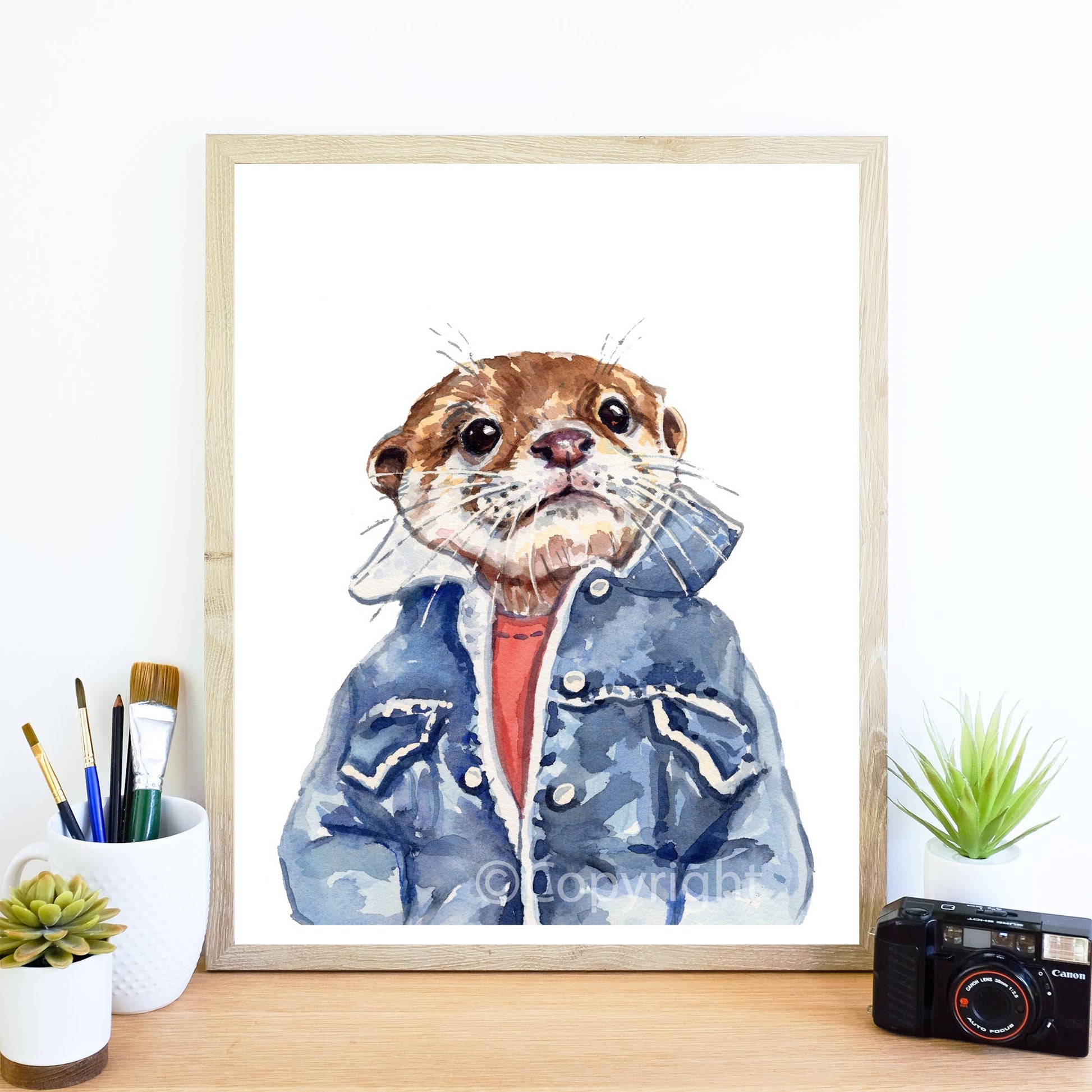 A framed watercolor art print of an otter wearing a blue denim jacket and orange shirt, displayed on a wooden desk with small plants and a camera.