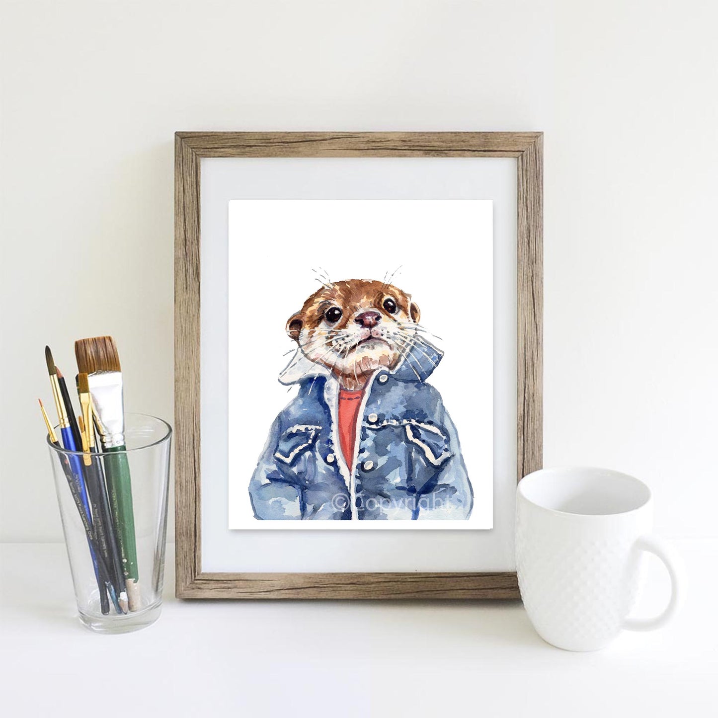 A framed watercolor art print of an otter wearing a blue jacket and red shirt, displayed on a white surface next to a glass of paintbrushes and a white ceramic mug.