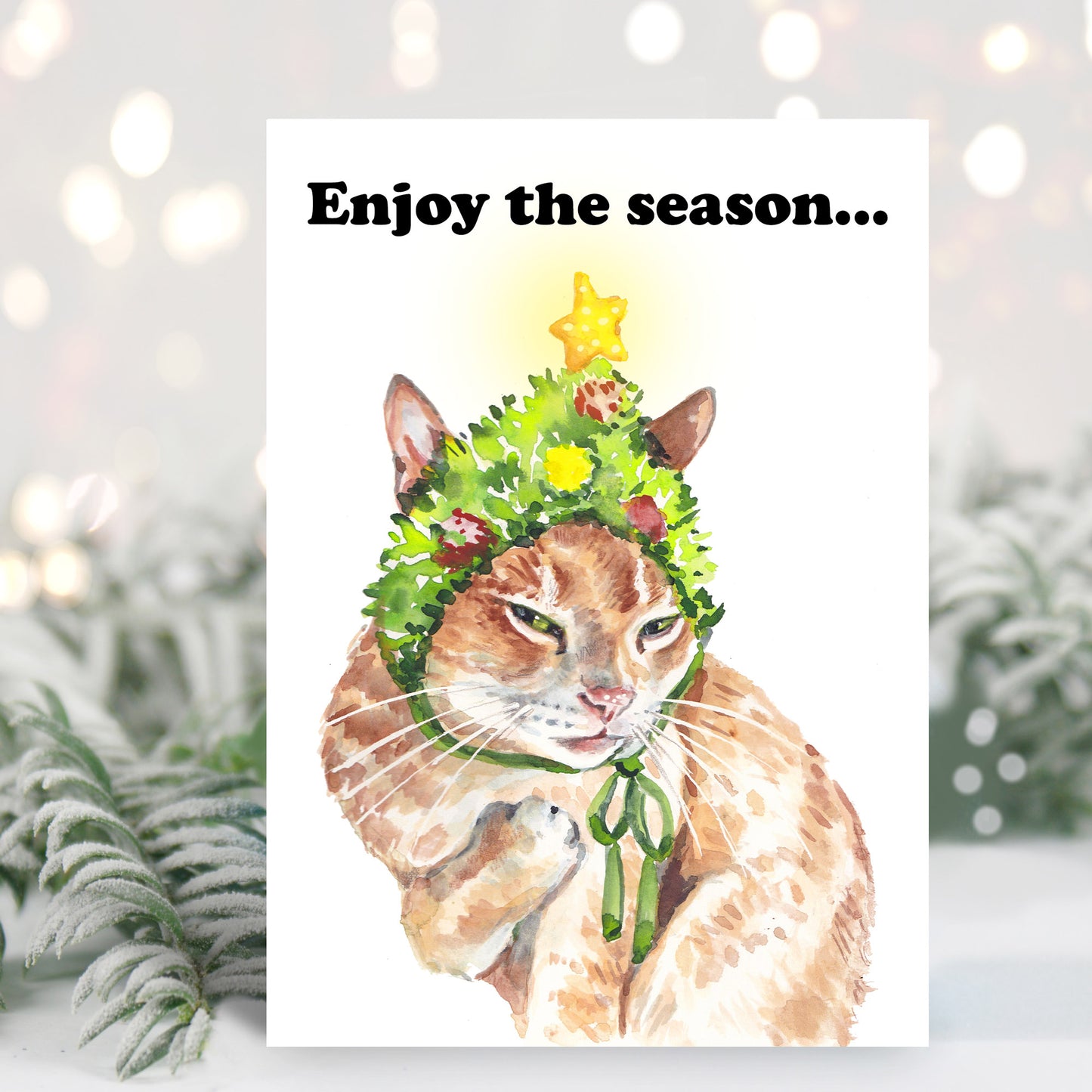 Christmas card featuring a grumpy orange cat wearing a silly Christmas tree hat. He plans to get even. 