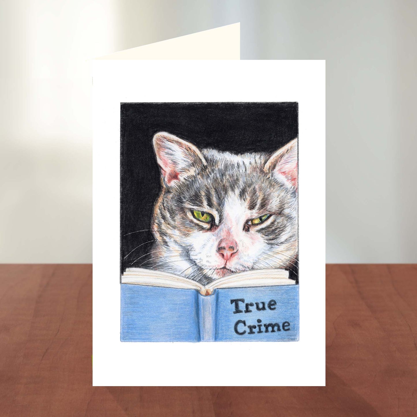 Greeting card featuring a coloured pencil drawing of a grey tabby cat reading a book about true crime. Art by Deidre Wicks