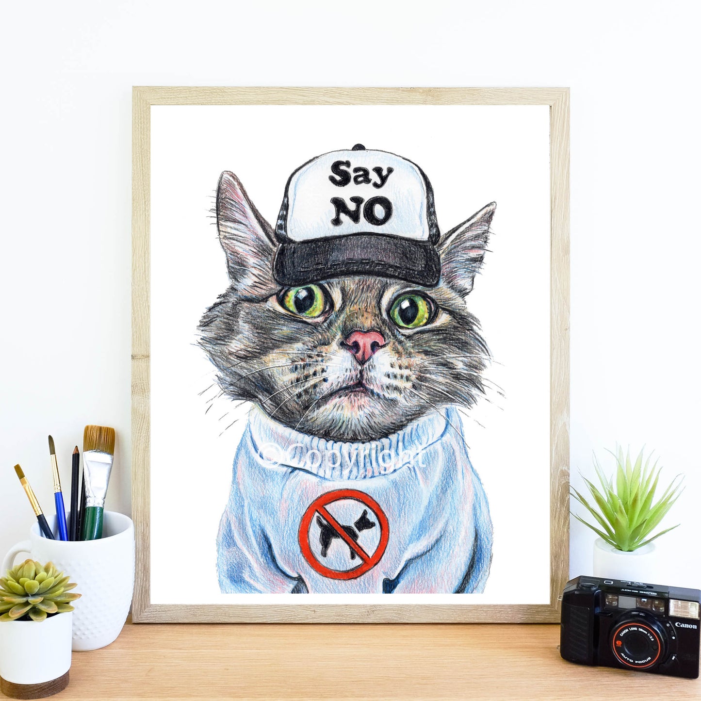 Framed artwork of a cat wearing a cap and a sweater with a no dogs sign on a desk.