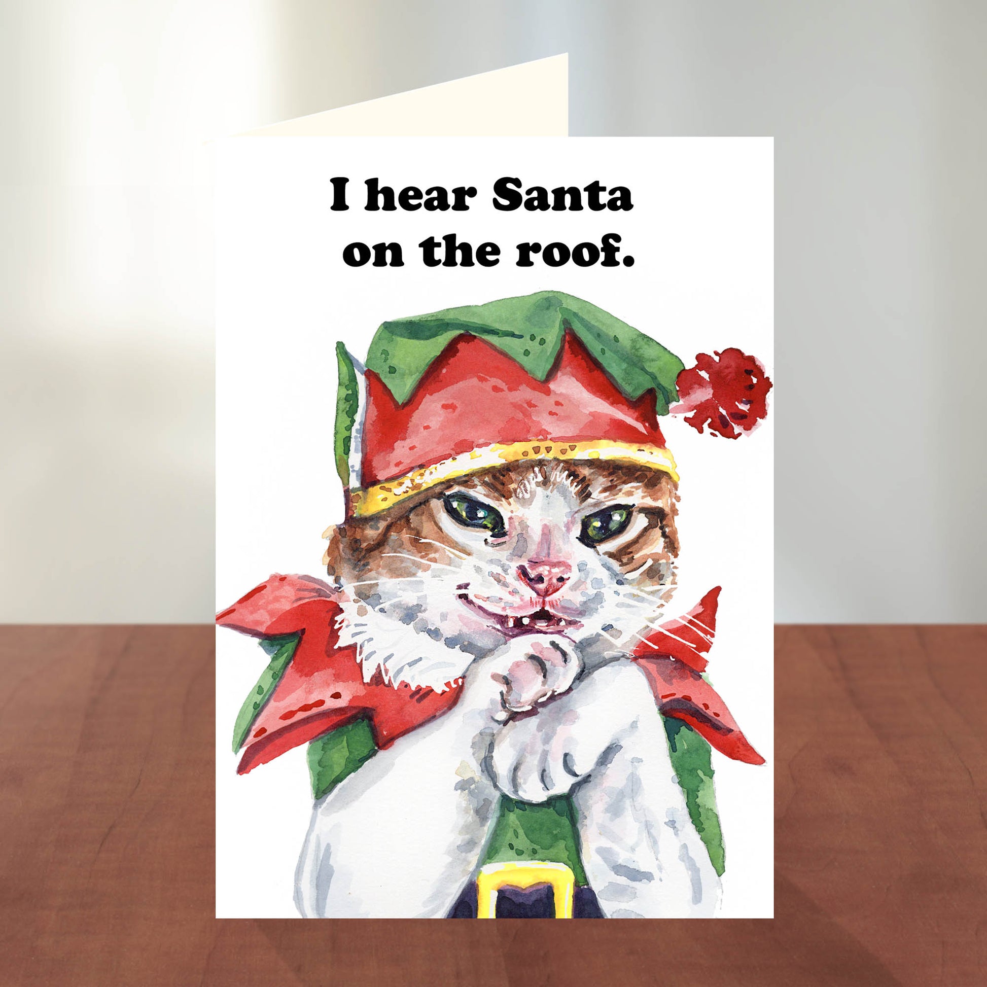 Cat in elf costume with text 'I hear Santa on the roof' on a white card.