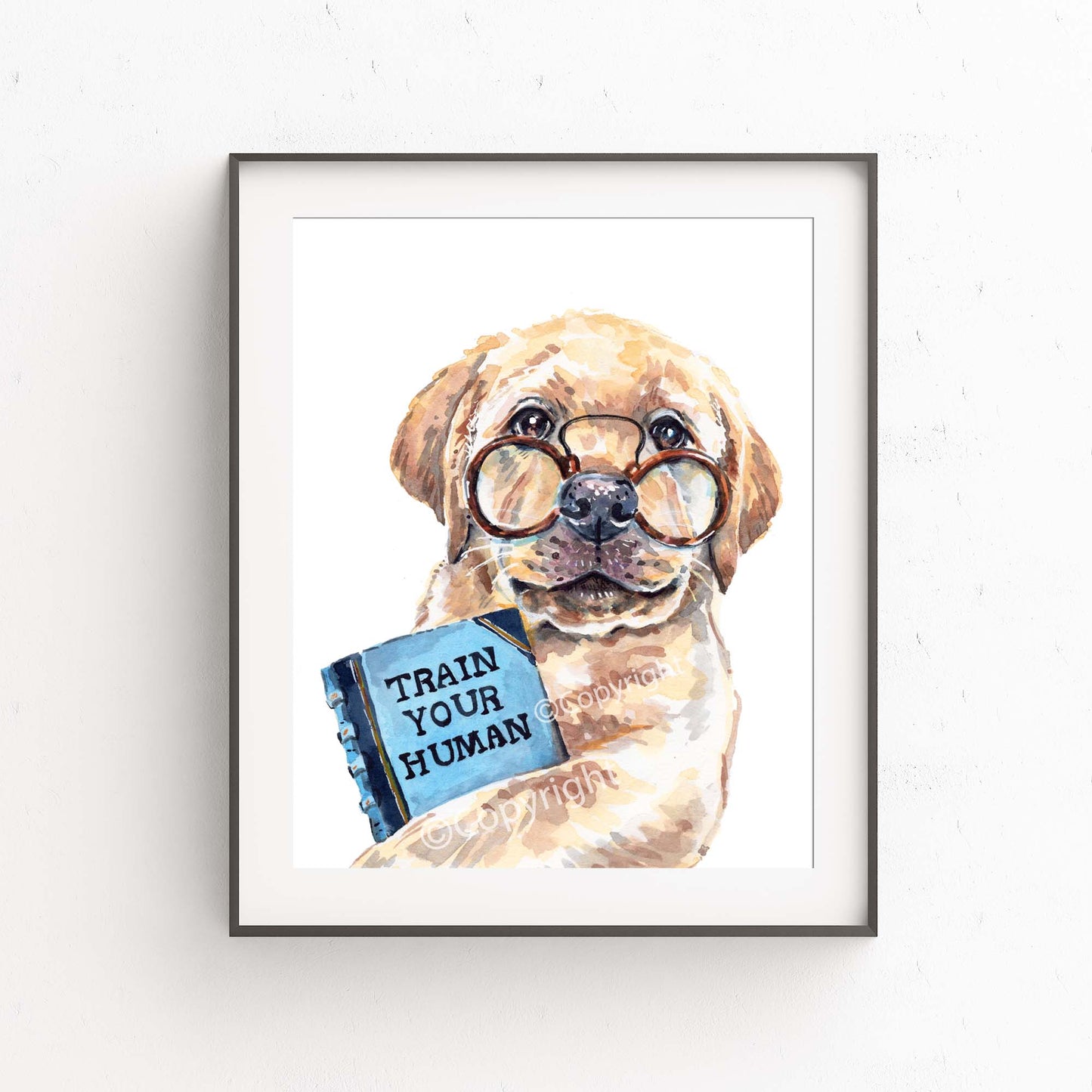 Framed artwork of a dog holding a book titled 'Train Your Human' on a white wall.