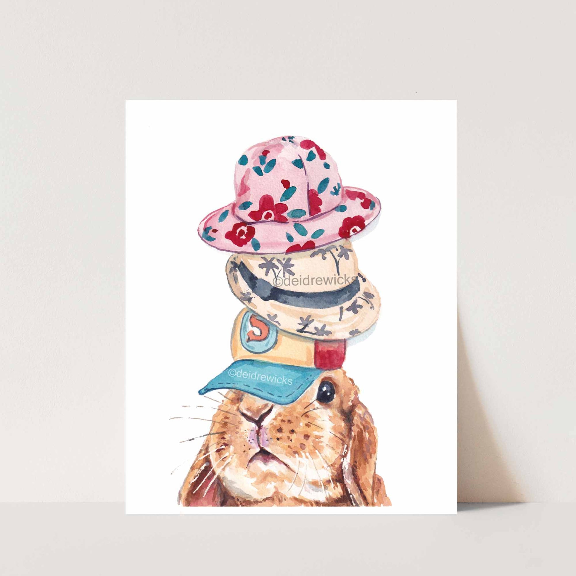 Watercolor print featuring a lop eared bunny rabbit wearing several hats. By Deidre Wicks